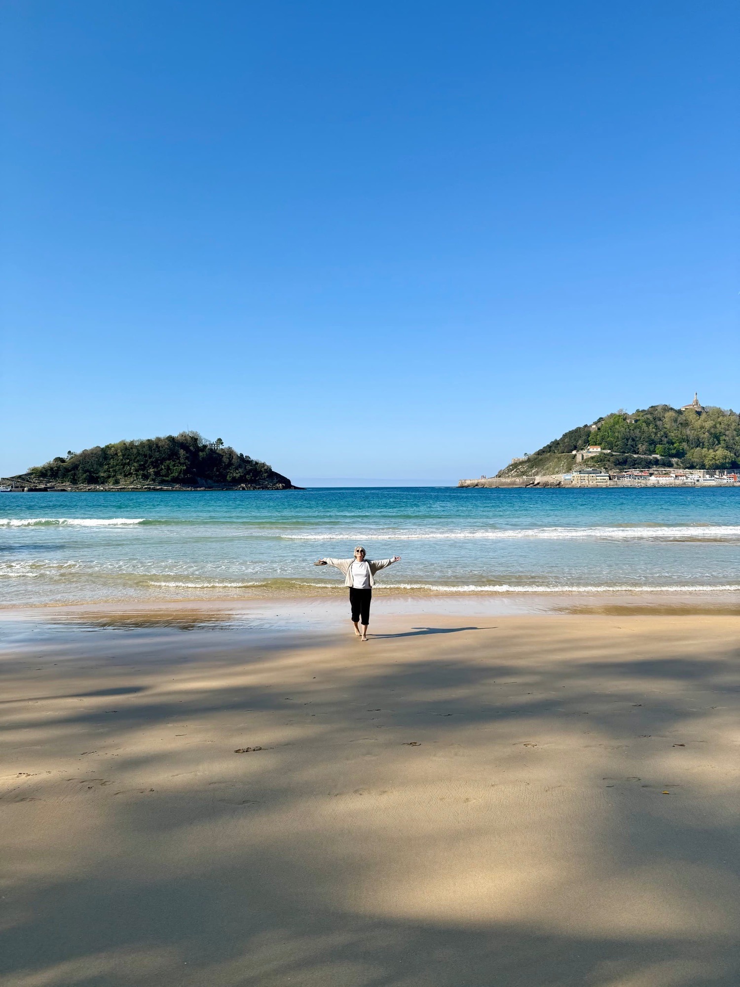 Blick über den Strand La Concha in Donostia-San Sebastián. Eine Person steht mit ausgebreiteten Armen am Wasser, im Hintergrund die Insel Santa Clara und die Bucht bei strahlend blauem Himmel.