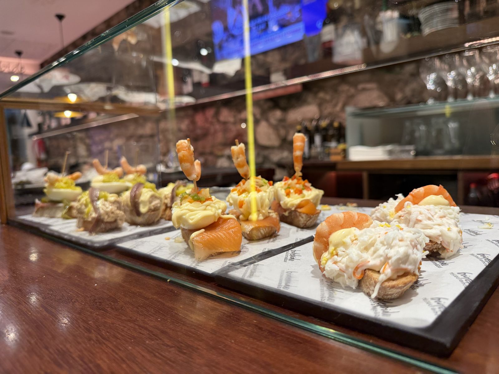 Glasvitrine auf der Holztheke im Bide Bide an der Legazpi Kalea in Donostia-San Sebastián mit Pintxos von Lachs, Garnelen, Ei und Anchoa, dahinter Natursteinwand und Weinregal