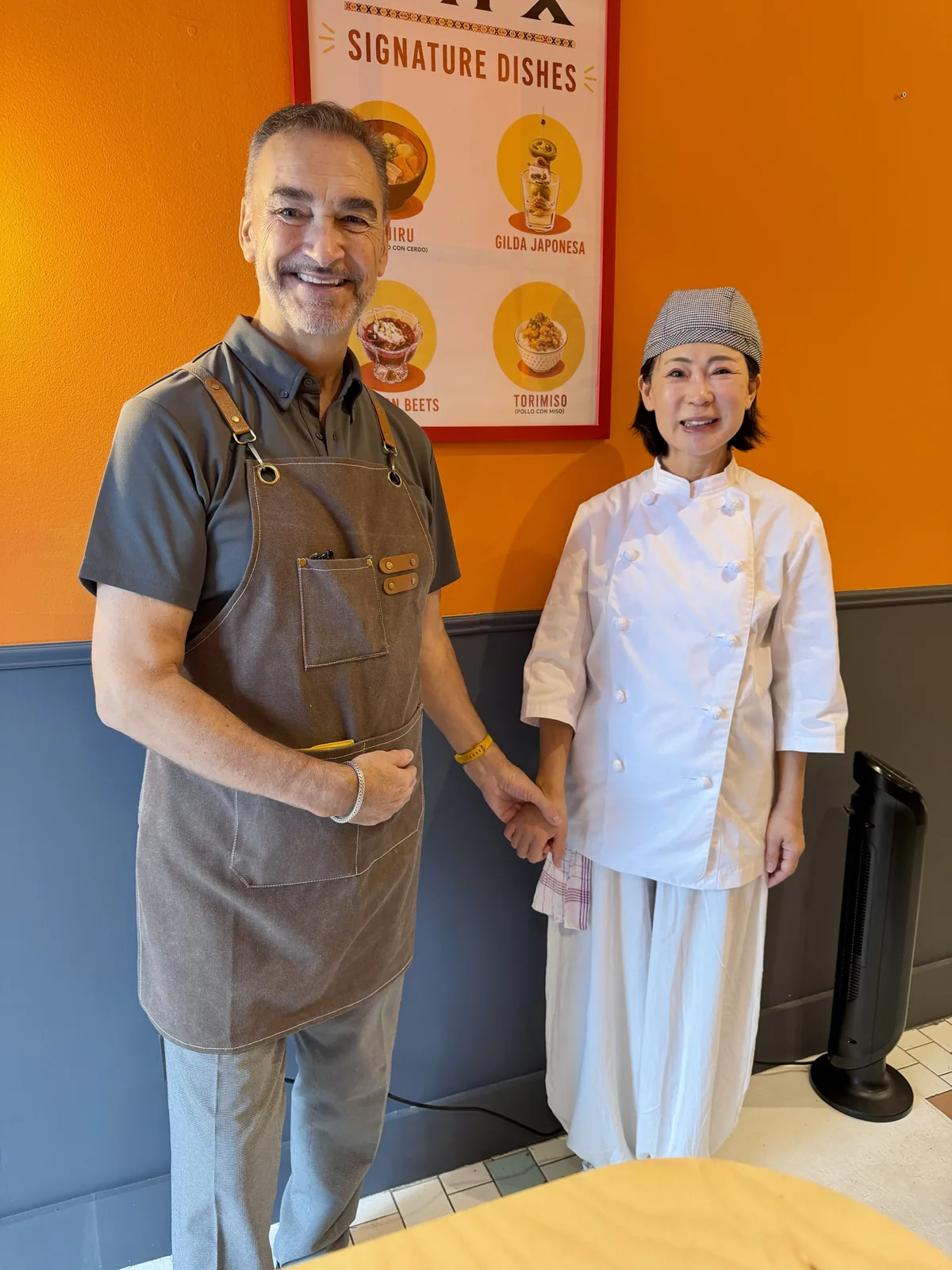 Köchin Emi in weißer Kochjacke und ihr Mann mit Schürze vor dem Plakat mit den Signature Dishes an der orangefarbenen Wand von Emi X