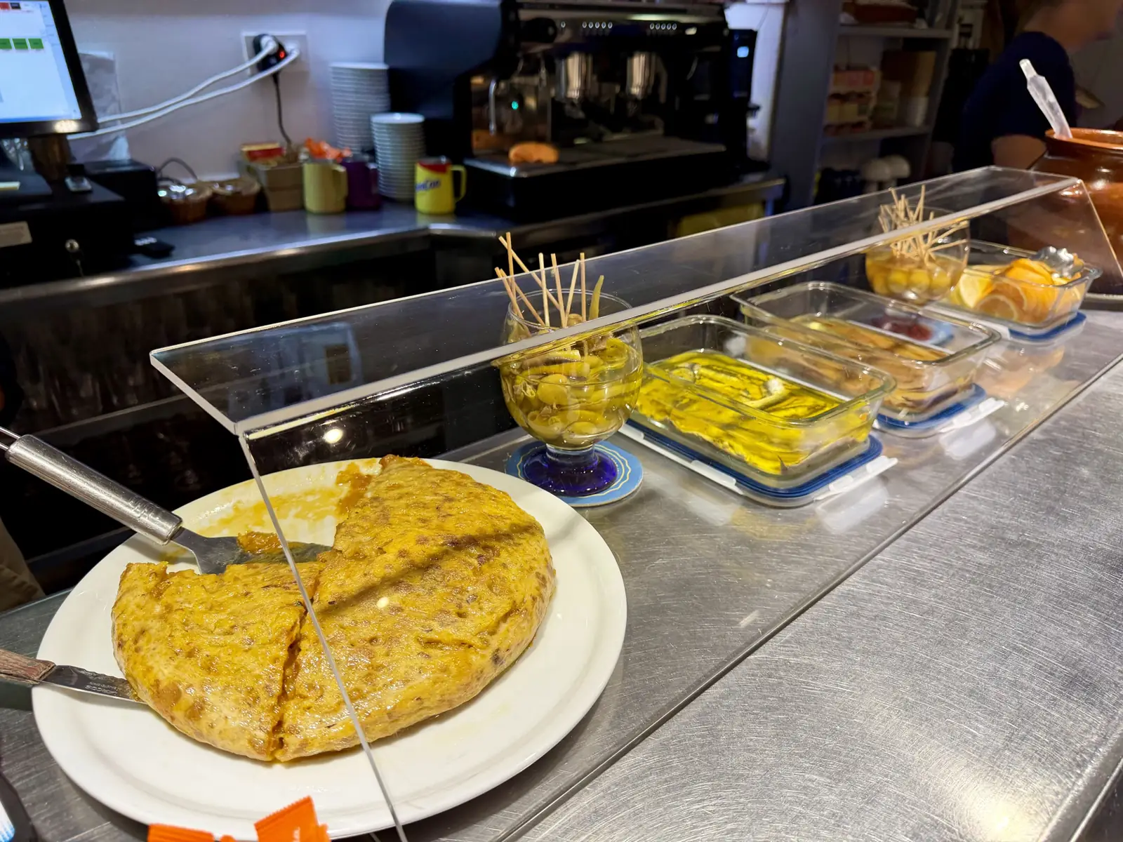Angeschnittene Tortilla de patata auf einem Teller an der Theke, daneben Gildas und Sardellen in Olivenöl hinter einer Glasscheibe